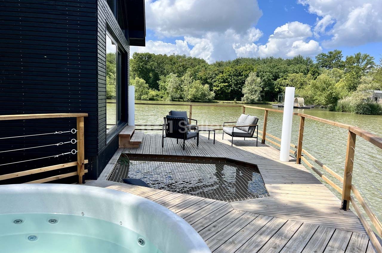 Terrasse en bois avec jacuzzi, fauteuils et filet suspendu, donnant sur un lac entouré de végétation.