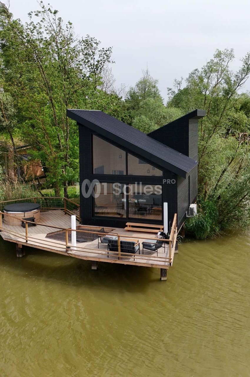 Maison moderne en bois noir avec terrasse sur pilotis, située au bord d'un plan d'eau entouré de végétation.