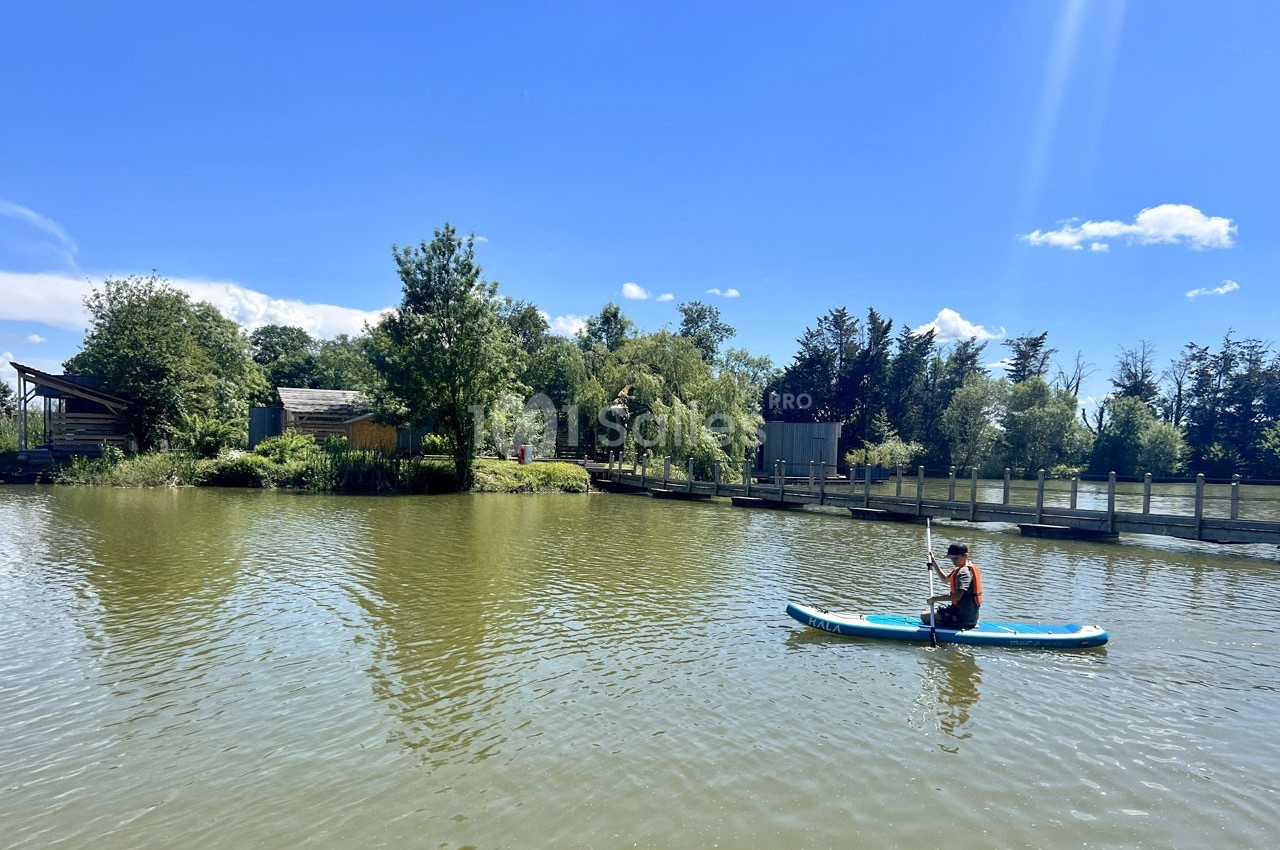 Une personne fait du paddle sur un lac entouré de végétation et de petites structures en bois sous un ciel bleu.