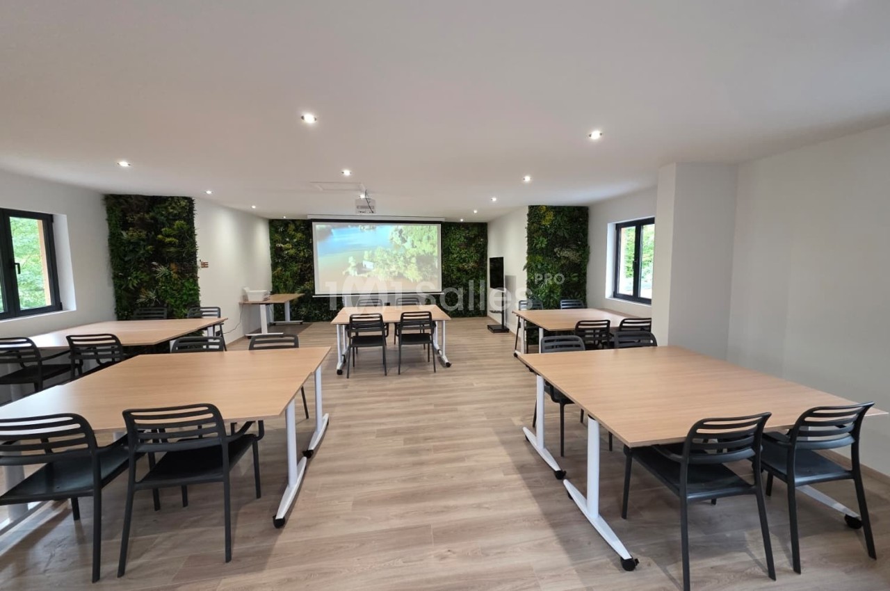 Salle de réunion lumineuse avec tables en bois, chaises noires, mur végétal et écran de projection.