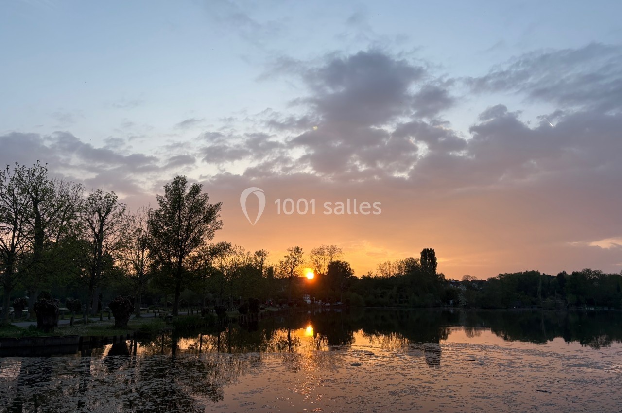 Coucher de soleil sur un lac entouré d'arbres, avec des reflets dorés sur l'eau et un ciel partiellement nuageux.