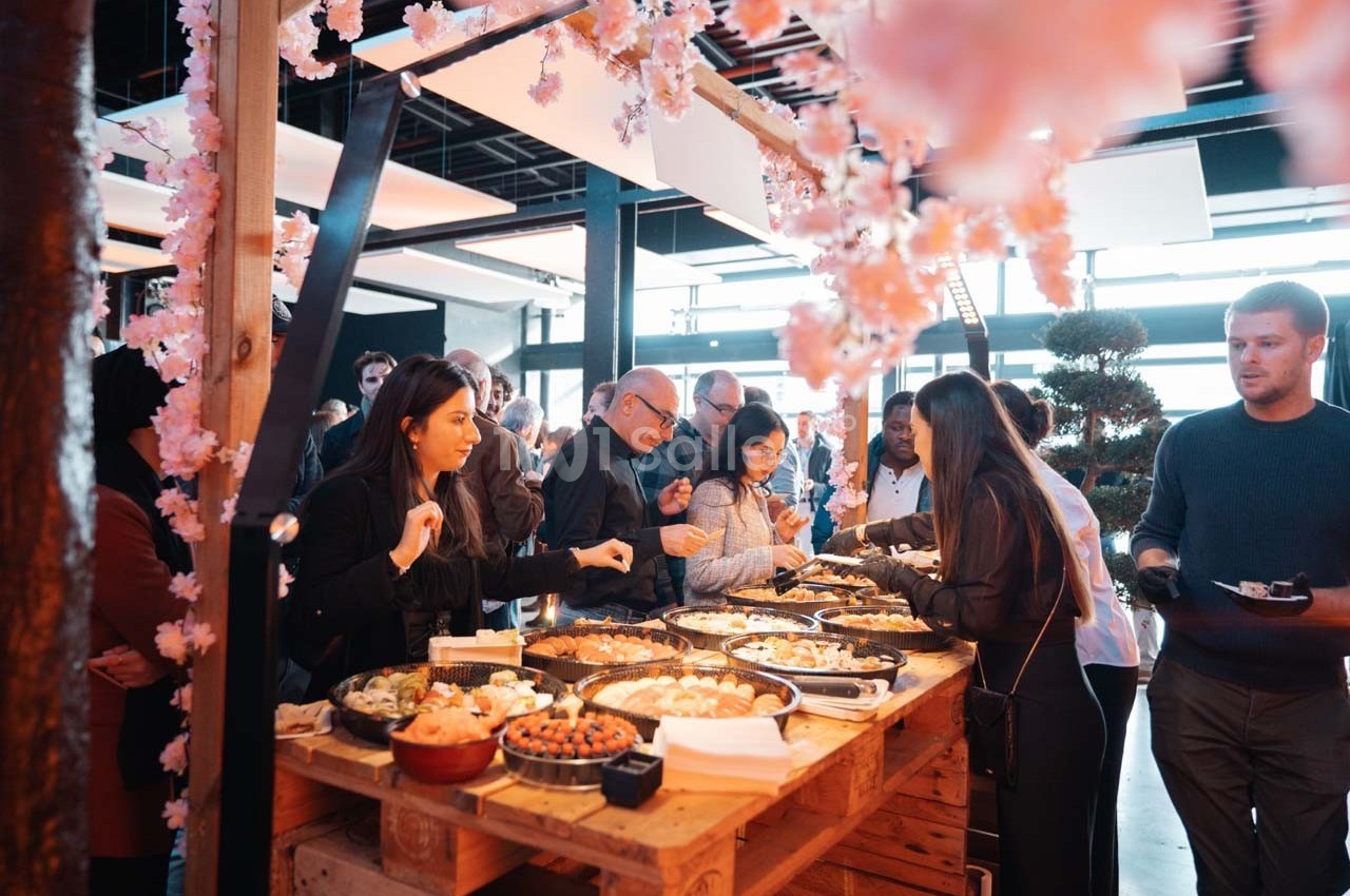 Des personnes se servent à un buffet garni de plats variés, sous des branches fleuries dans un espace lumineux.
