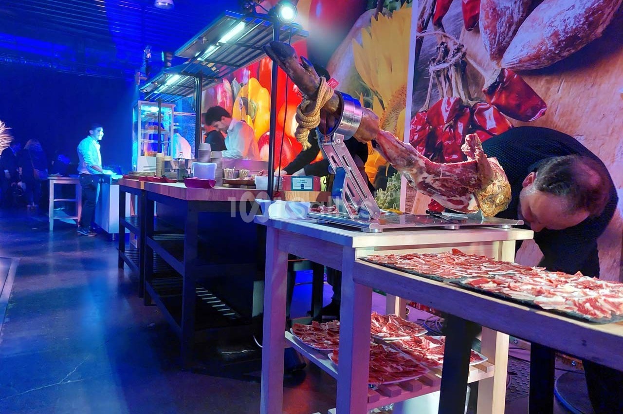 Découpe de jambon sur un stand de restauration dans un espace éclairé, avec des tables et des décorations colorées en…