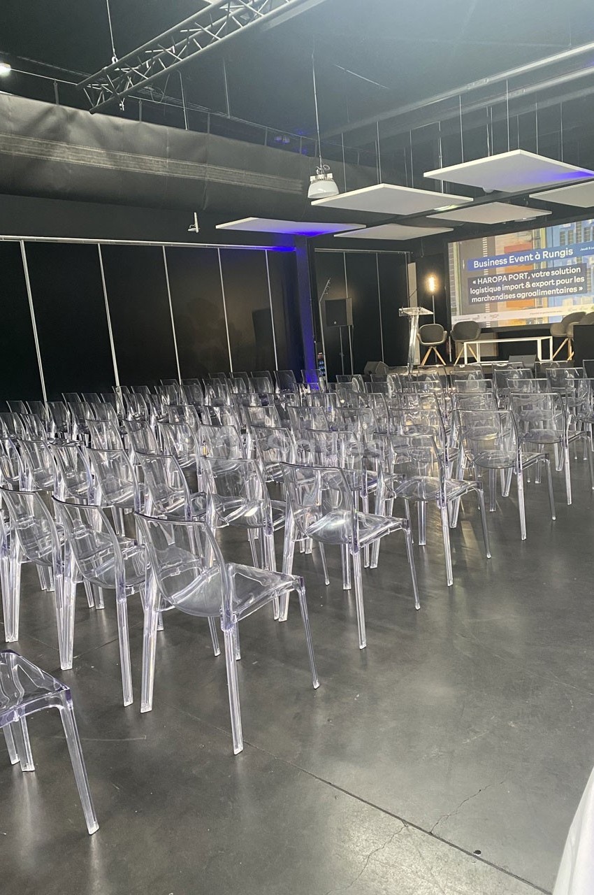 Salle de conférence avec rangées de chaises transparentes alignées devant une scène équipée d'un écran de présentation.