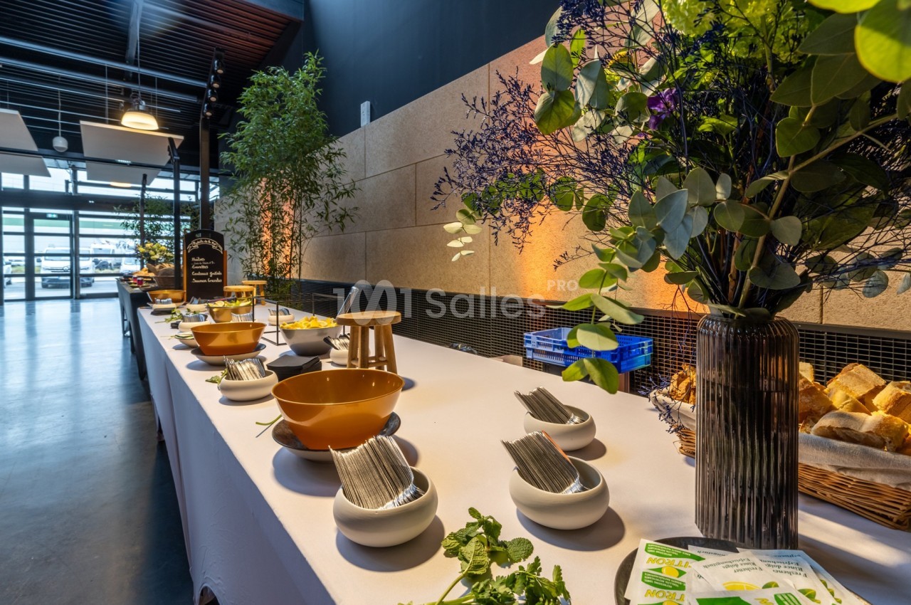 Buffet avec bols, couverts, pains et décorations florales sur une table blanche dans une salle lumineuse.