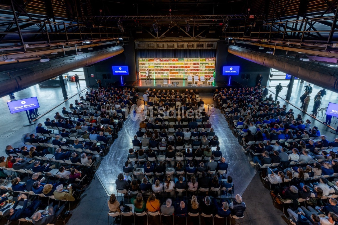 Salle comble avec un public assis face à une scène, entourée d'écrans et d'étagères colorées en arrière-plan.