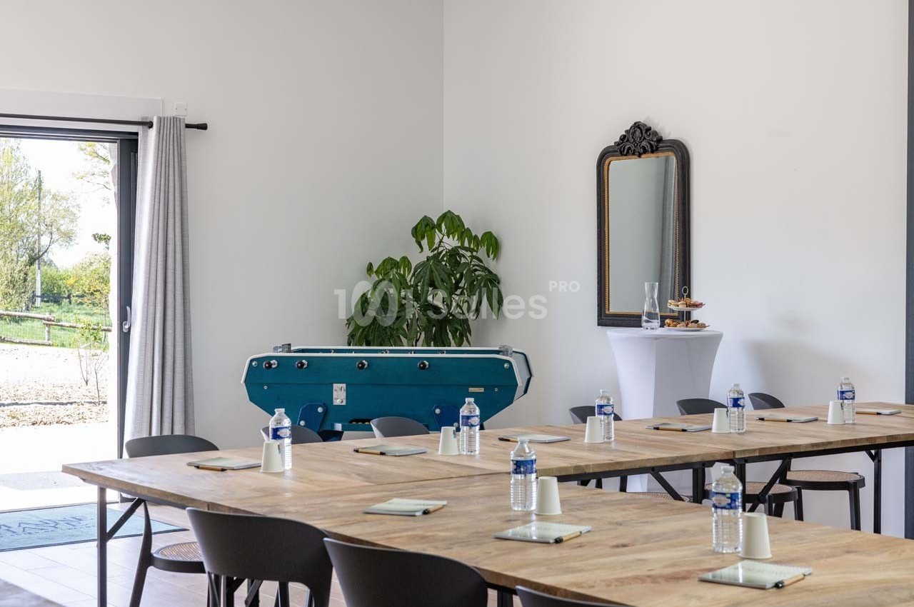 Salle lumineuse aménagée avec des tables en bois, des chaises, un baby-foot, un miroir et des bouteilles d'eau.