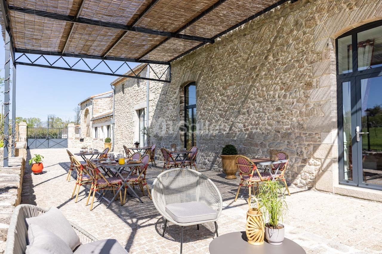 Terrasse ensoleillée avec tables et chaises en rotin, adossée à un bâtiment en pierre avec grandes ouvertures vitrées.