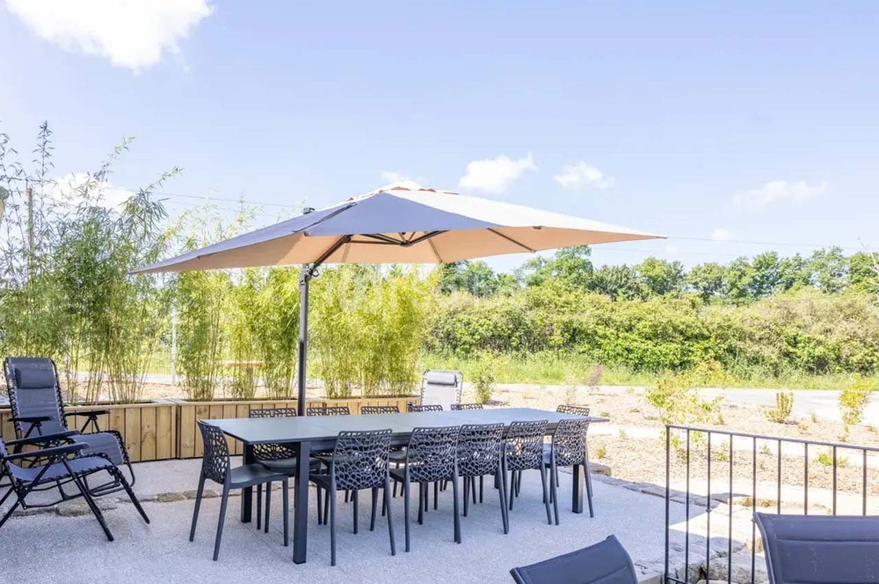 Table de jardin avec chaises noires et grand parasol beige, située sur une terrasse ensoleillée entourée de verdure.