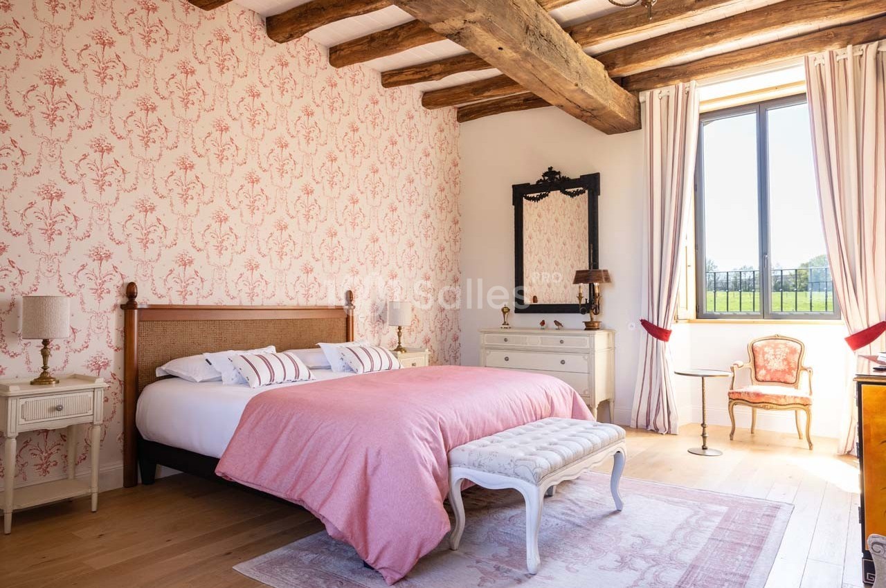 Chambre lumineuse avec lit double, murs ornés de papier peint floral, poutres apparentes et vue sur un jardin.