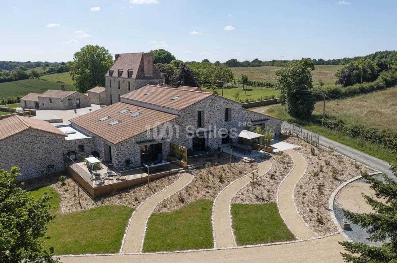 Vue aérienne d'un bâtiment en pierre rénové avec toits en tuiles, entouré de chemins et de verdure en milieu rural.