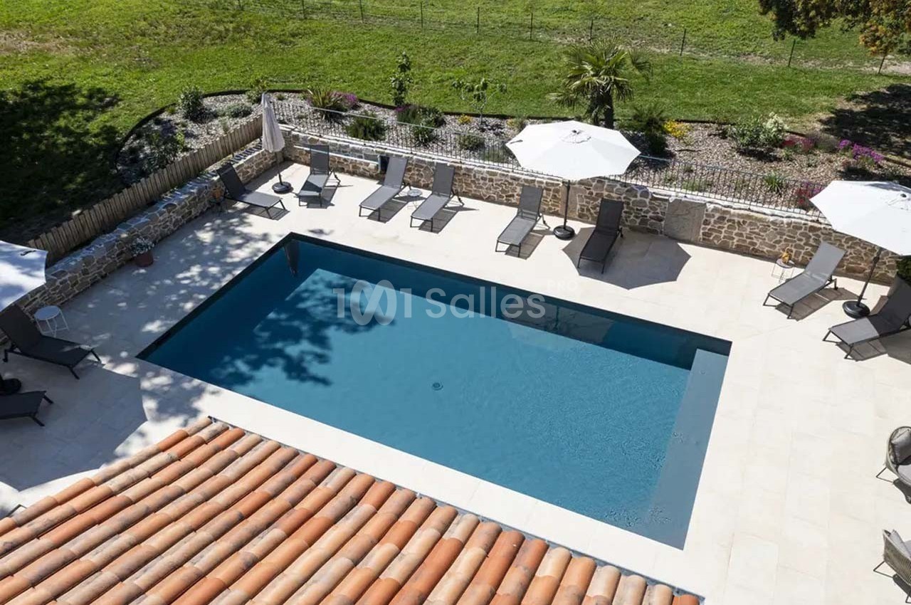 Vue aérienne d'une piscine rectangulaire entourée de transats, parasols et d'un jardin clos par un muret en pierre.