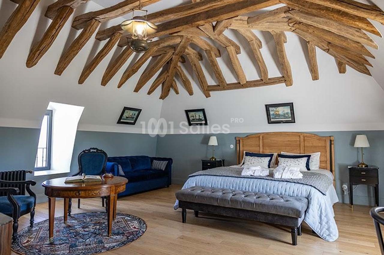Chambre spacieuse avec poutres apparentes, lit double, canapé bleu, table ronde et décoration classique.