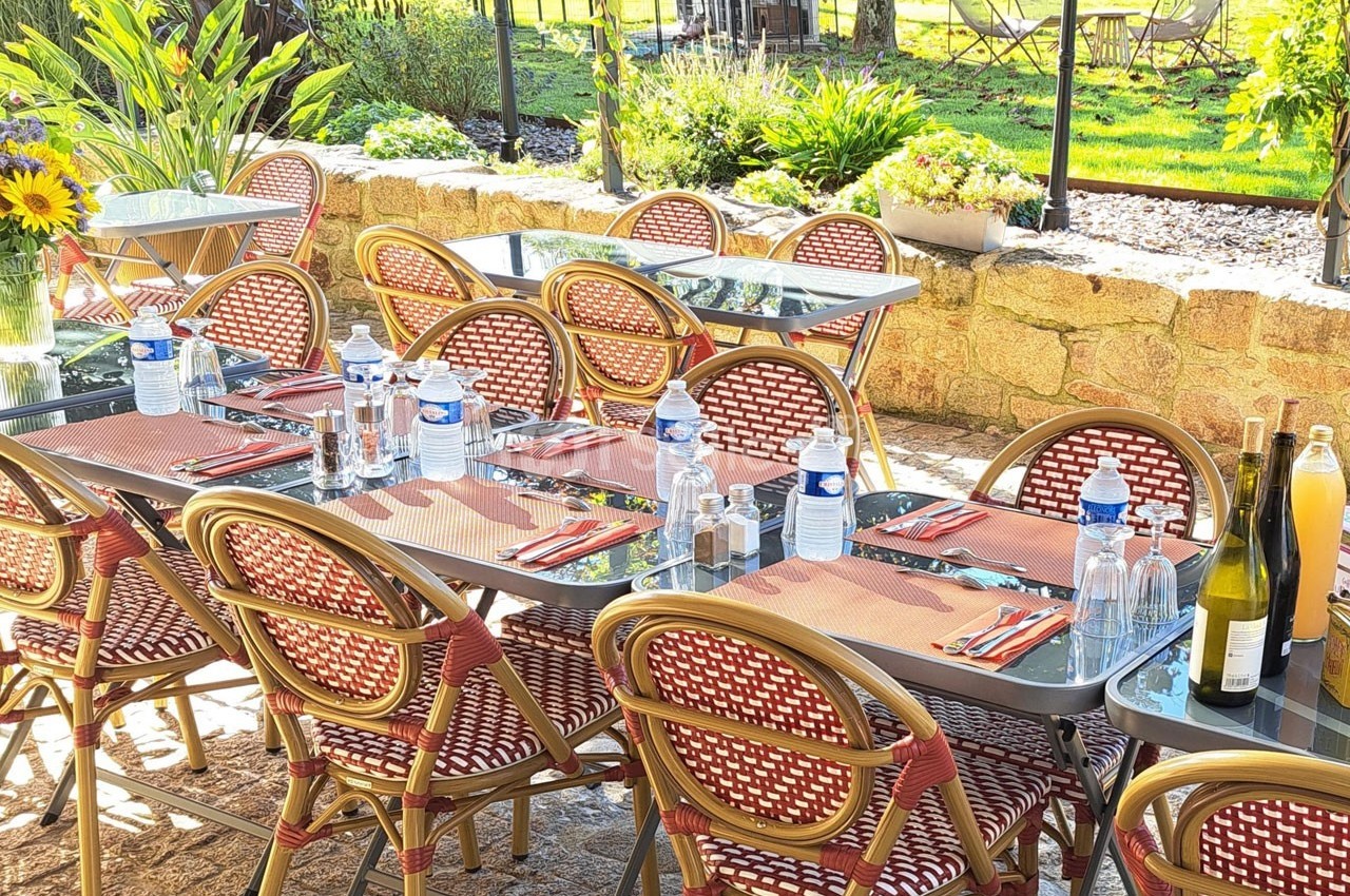 Terrasse ensoleillée avec tables dressées, chaises en osier, bouteilles d'eau et de vin, vue sur un jardin verdoyant.