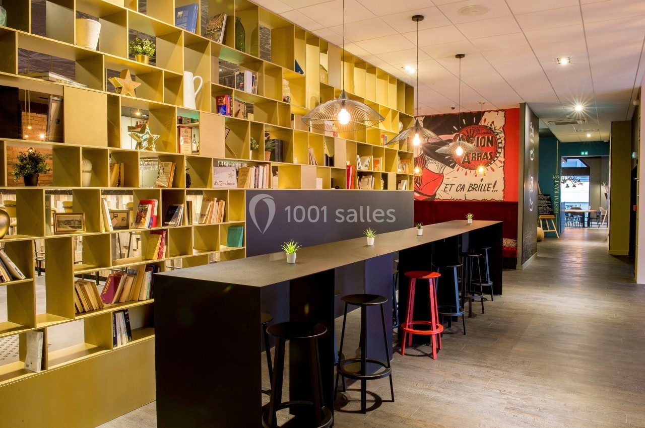 Espace intérieur moderne avec étagères remplies de livres, table haute, tabourets et éclairage suspendu.