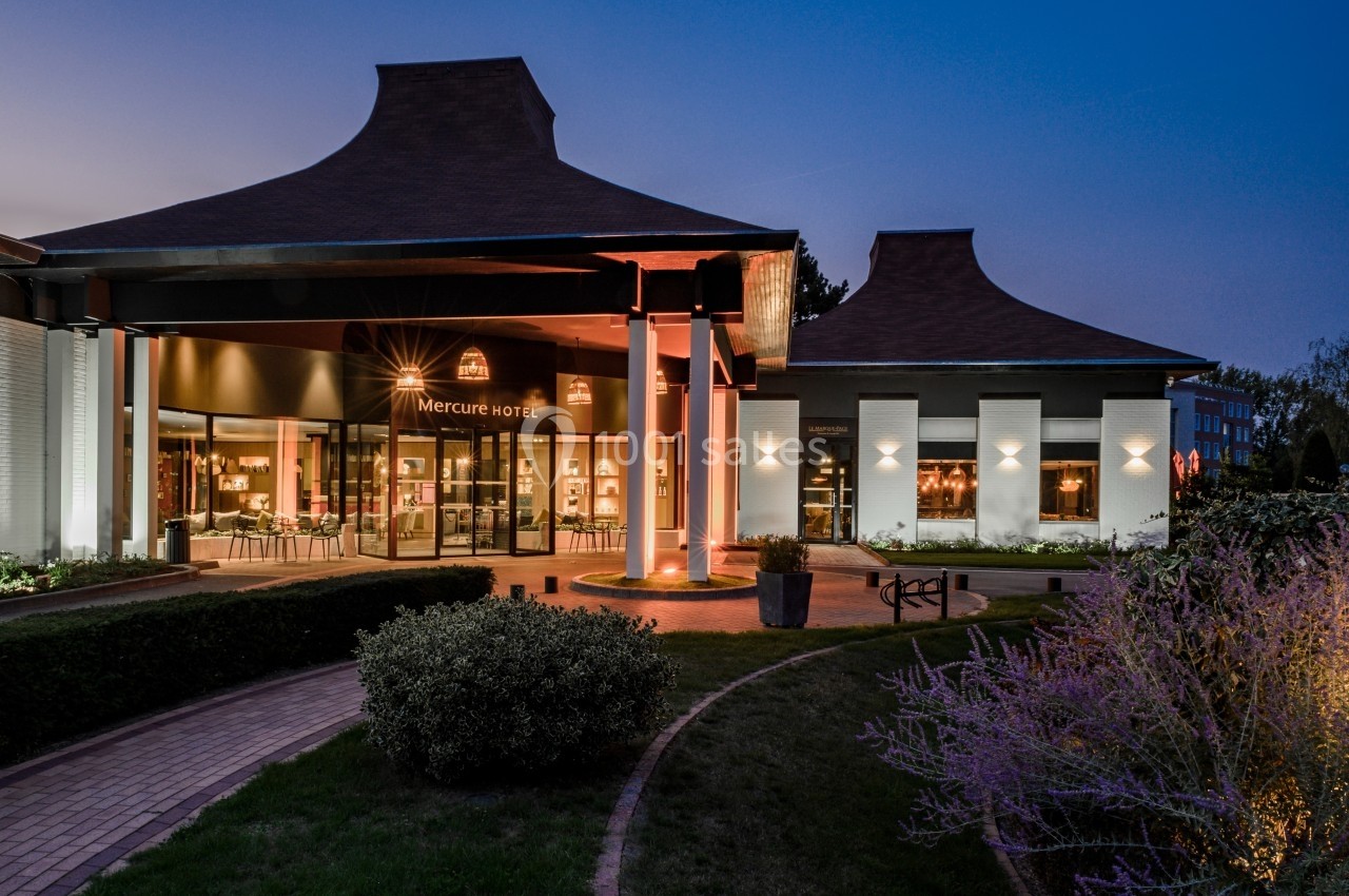 Entrée d'un hôtel moderne éclairée en soirée, avec jardin aménagé et chemin pavé au premier plan.