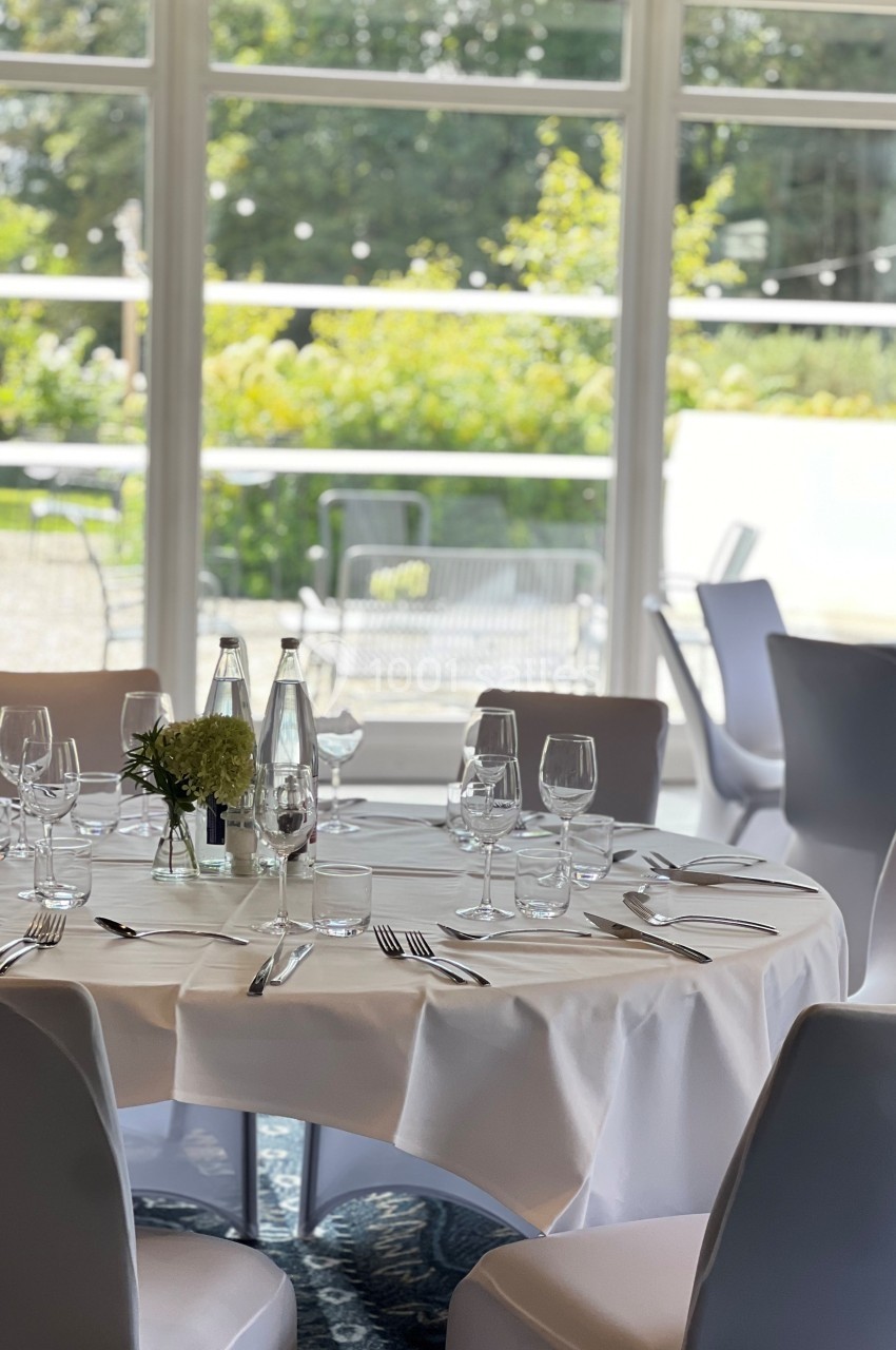 Table dressée avec nappe blanche, verres, couverts et centre de table floral, devant une baie vitrée donnant sur un jardin.