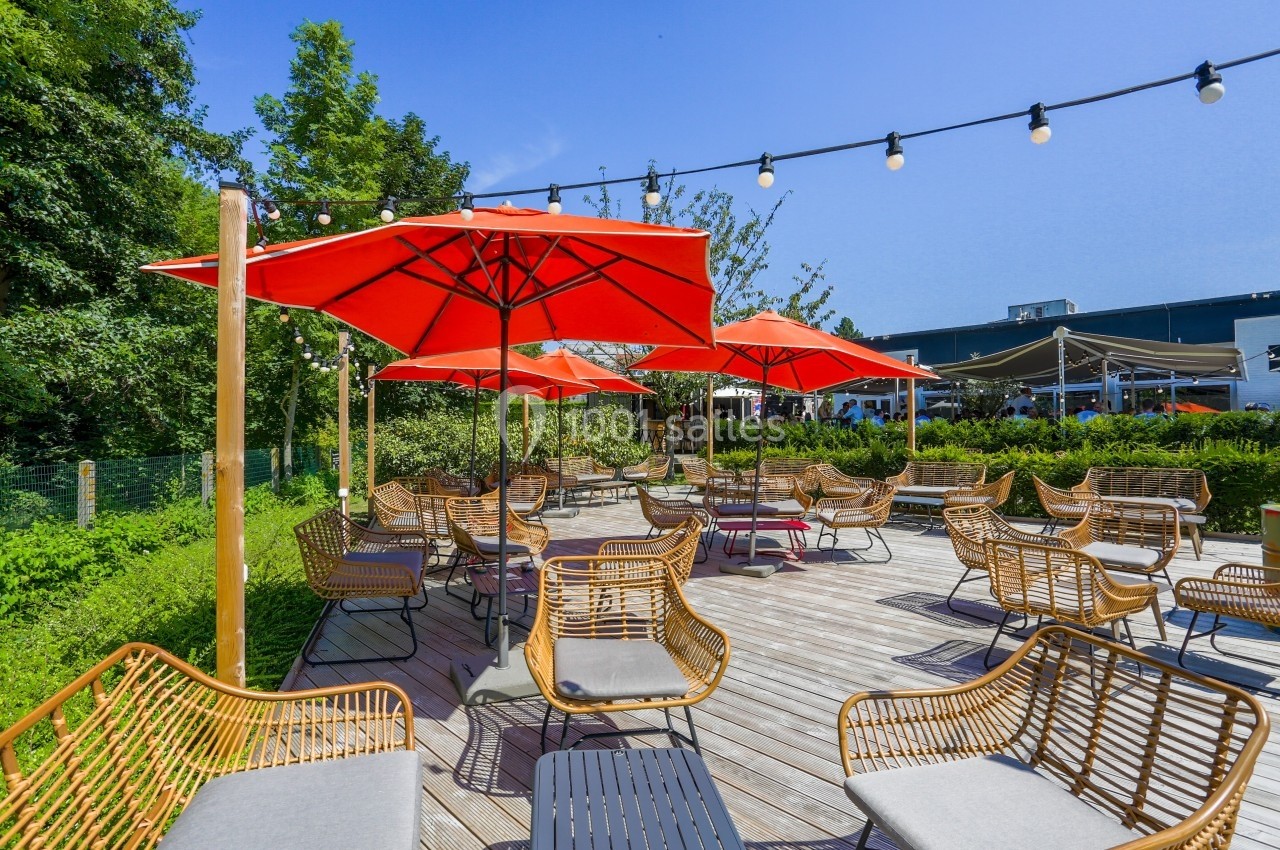 Terrasse en bois avec des fauteuils en rotin, des parasols rouges et des guirlandes lumineuses, entourée de verdure.