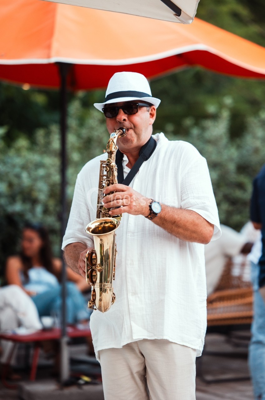 Un homme en chemise blanche et chapeau joue du saxophone sous un parasol rouge en extérieur.