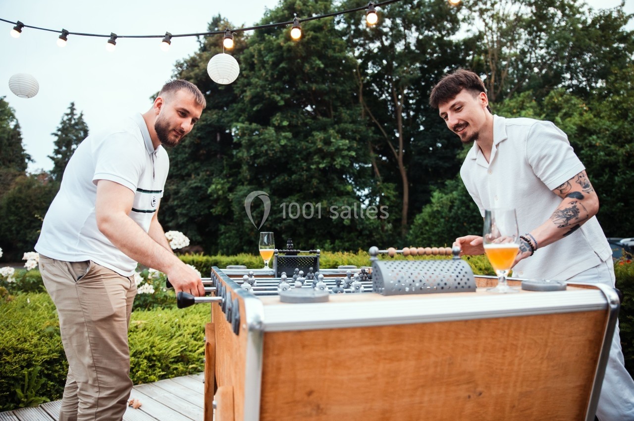 Deux hommes jouent au baby-foot en extérieur, entourés de verdure et de guirlandes lumineuses.