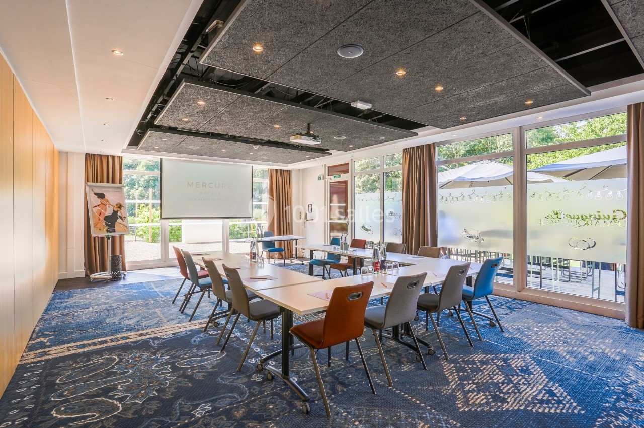 Salle de réunion lumineuse avec tables disposées en U, chaises colorées et grandes baies vitrées donnant sur une terrasse.