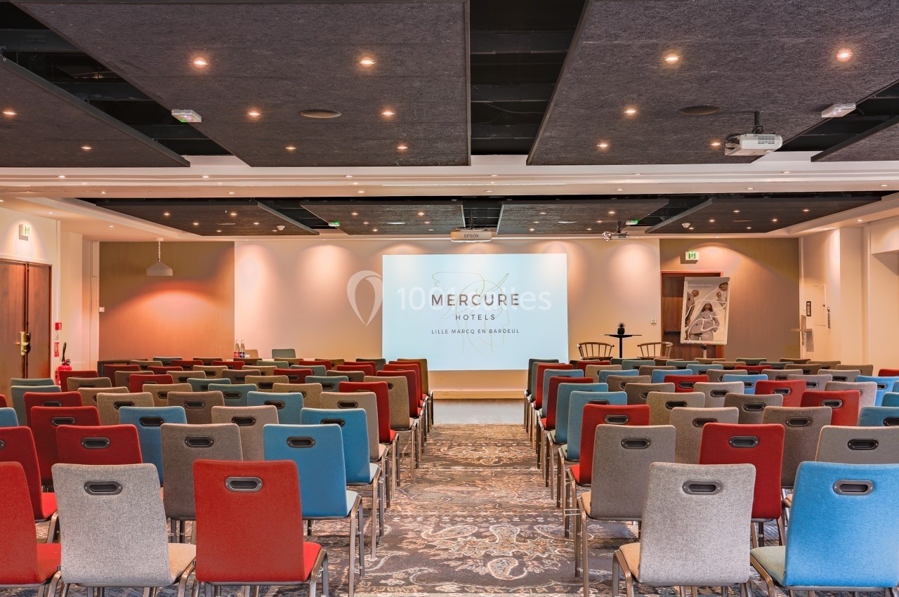 Salle de conférence avec des rangées de chaises colorées face à un écran de projection allumé.