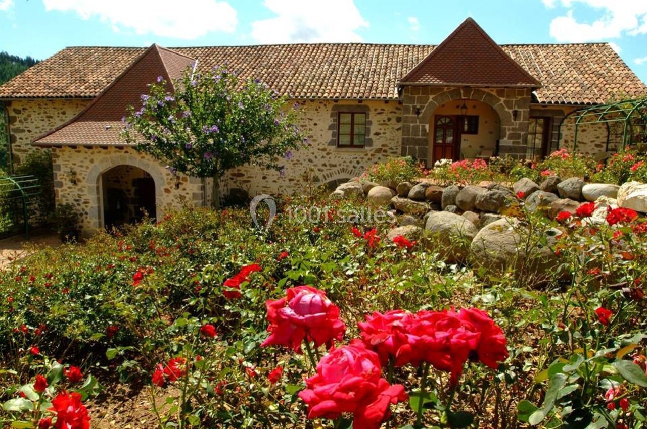 Maison en pierre avec toit en tuiles, entourée de rosiers rouges et d'un arbre en fleurs sous un ciel bleu.