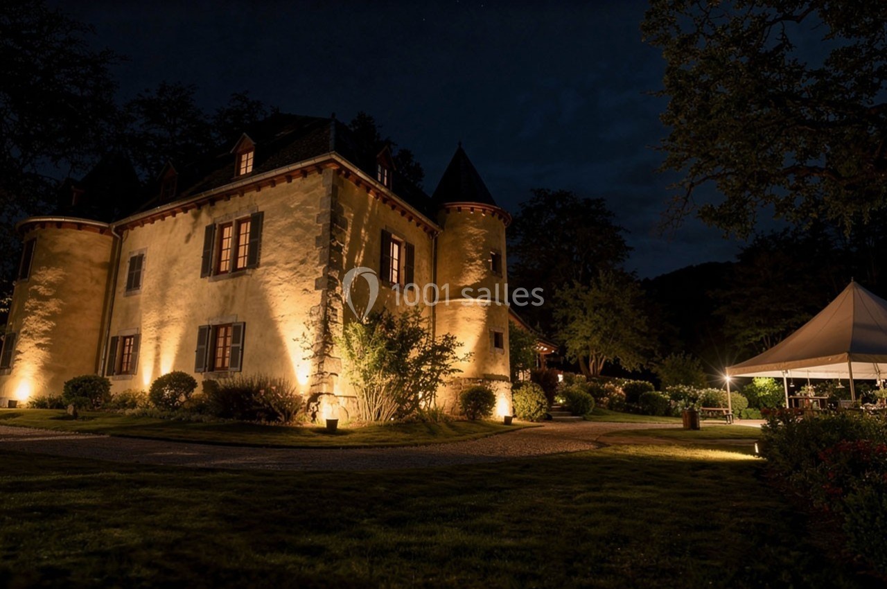 Château éclairé de nuit avec un jardin aménagé et une tente blanche éclairée sur la droite.