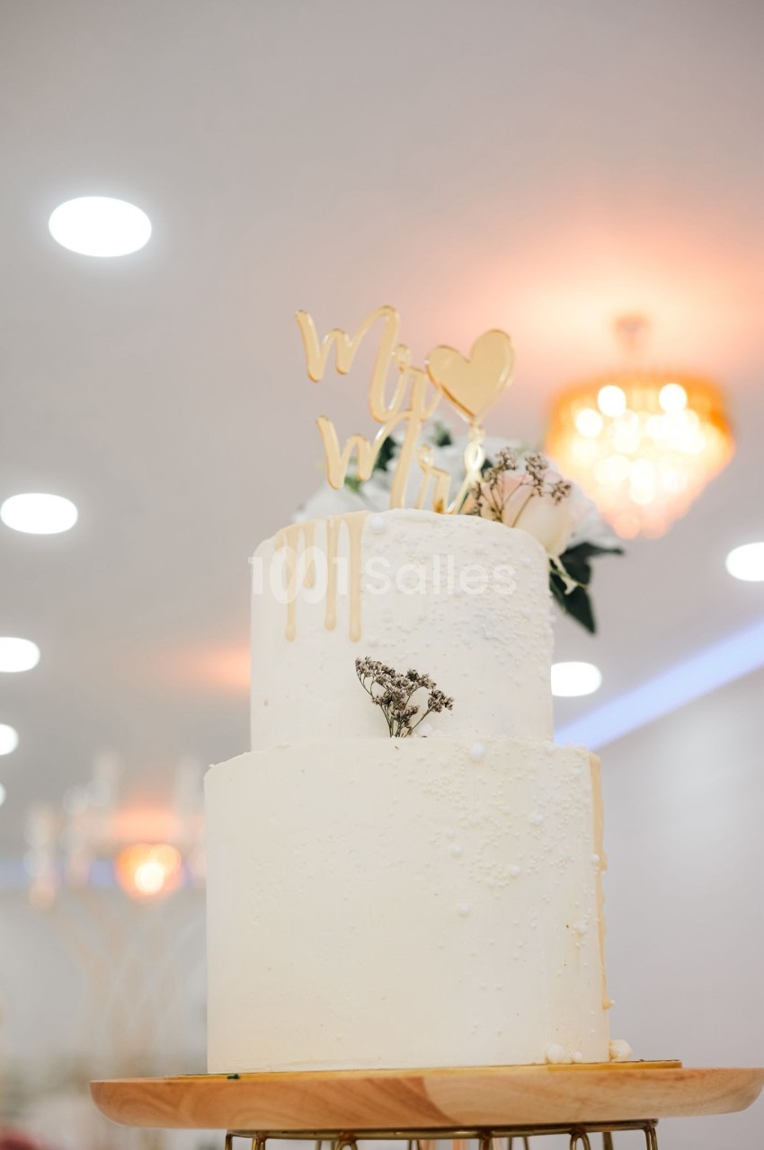 Gâteau de mariage à deux étages décoré de fleurs séchées et d'un topper doré, sur un support en bois.