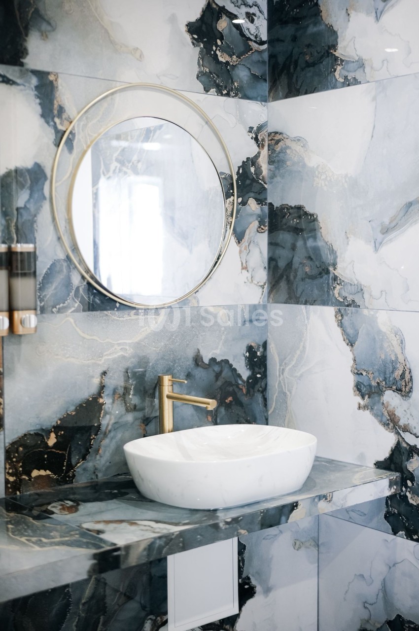 Salle de bain moderne avec un lavabo en céramique blanche, un robinet doré et un miroir rond sur un mur en marbre.