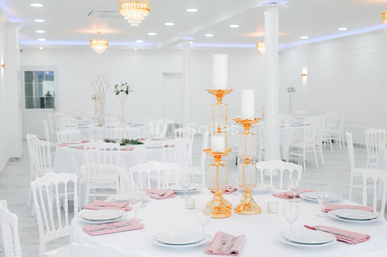 Salle de réception lumineuse avec tables rondes dressées, nappes blanches, serviettes roses et chandeliers dorés.