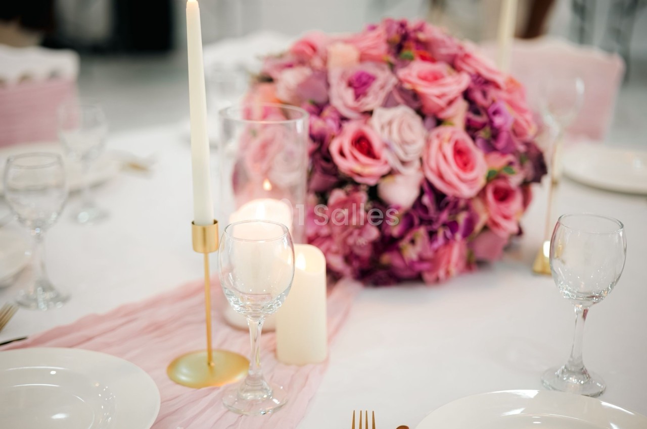 Centre de table avec bouquet de roses roses et violettes, bougies allumées et vaisselle sur une nappe blanche.