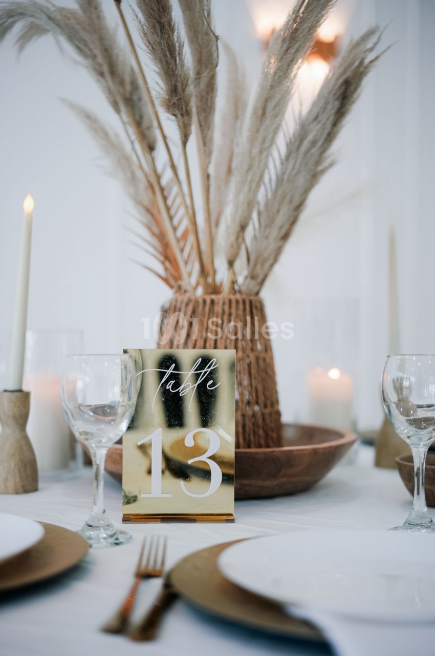 Table décorée avec des assiettes blanches, bougies, pampas et un numéro de table 13 sur un support doré.