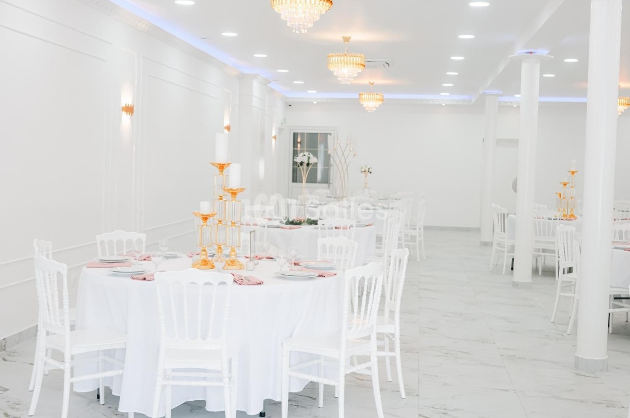 Salle de réception lumineuse avec tables rondes dressées, chaises blanches et chandeliers dorés.