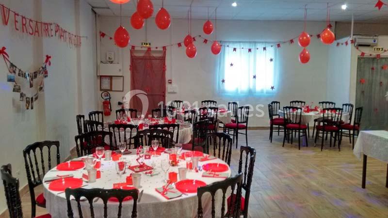Salle décorée pour une fête, avec des ballons rouges, des tables dressées et des guirlandes suspendues.