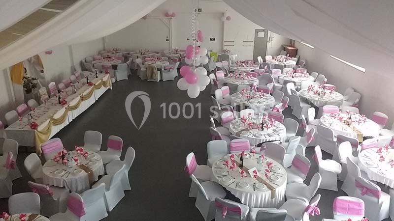 Salle de réception décorée avec des tables rondes, nappes blanches et chaises ornées de rubans roses.