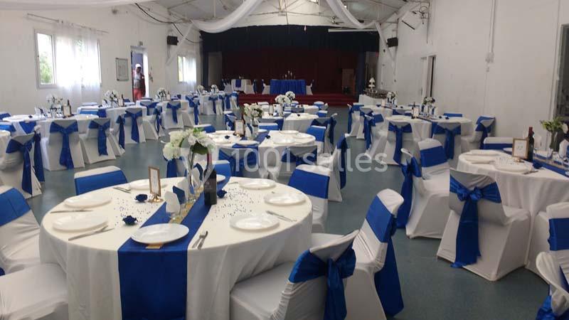 Salle décorée pour un événement, avec des tables rondes dressées et des chaises blanches ornées de nœuds bleus.