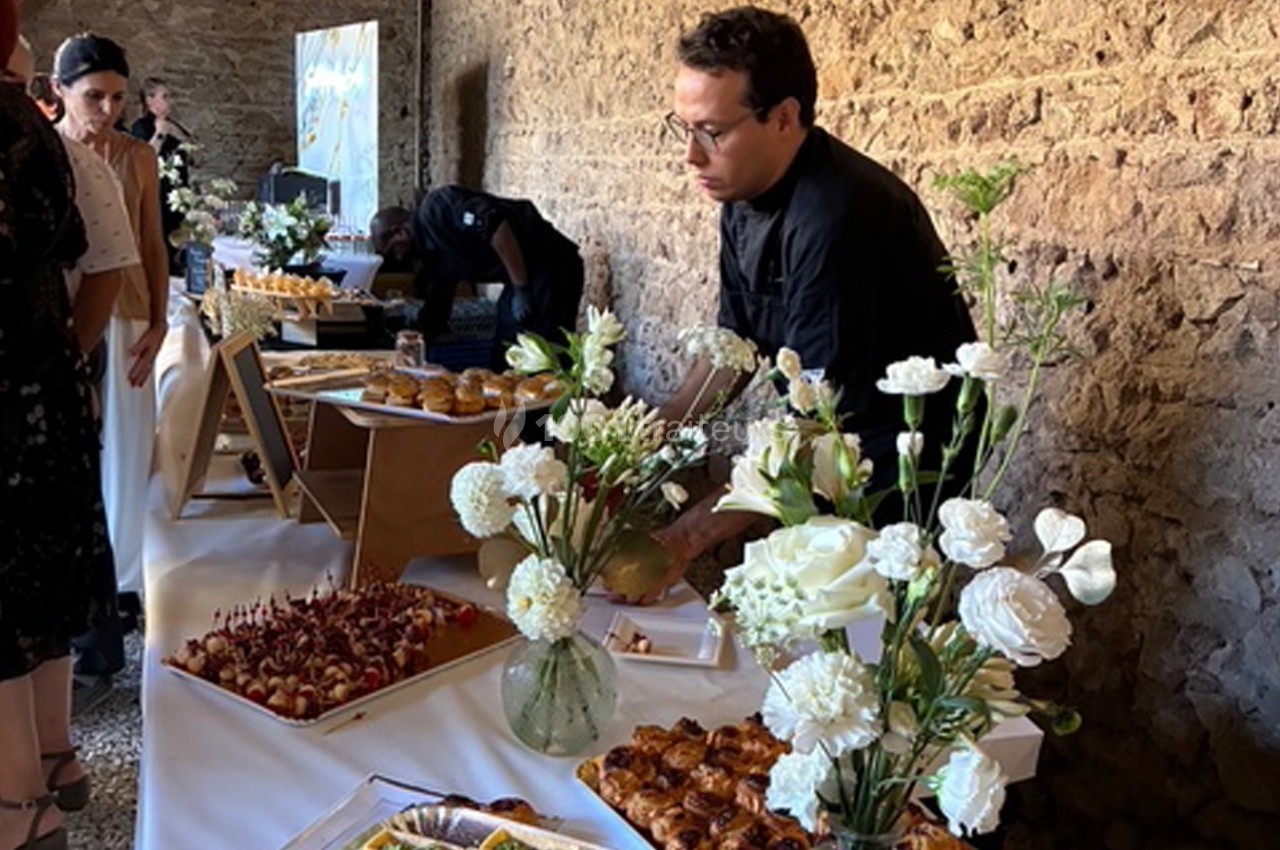 Un traiteur dispose des plats sur une table garnie de fleurs blanches dans une salle en pierre.