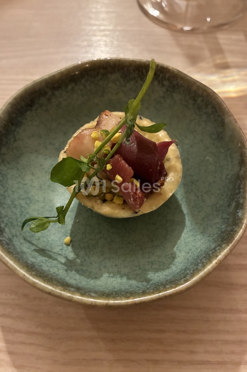 Tartelette garnie de viande séchée, herbes fraîches et éclats de pistache, servie dans une assiette en céramique bleue.