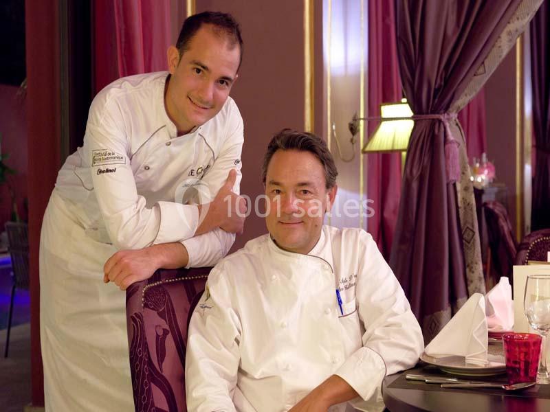 Deux chefs en tenue blanche posent dans un restaurant élégant avec des rideaux violets en arrière-plan.