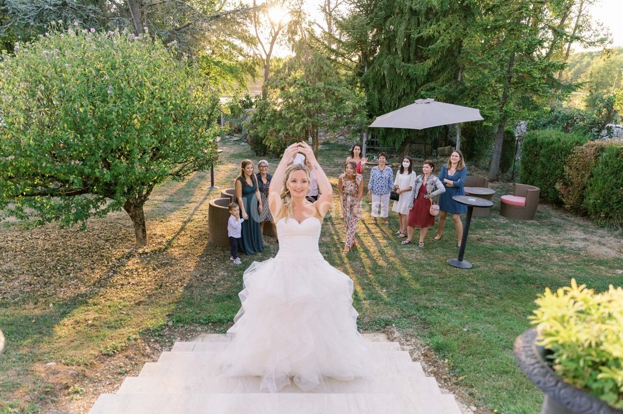 Une mariée lance son bouquet depuis des marches, entourée d'invités dans un jardin ensoleillé.