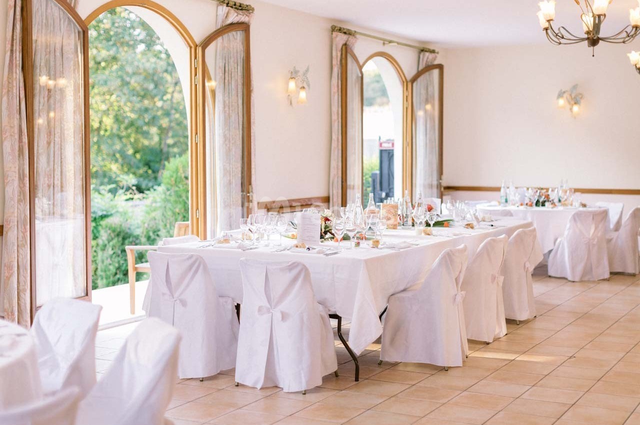 Salle lumineuse avec grandes fenêtres ouvertes, tables dressées avec nappes blanches et chaises décorées de nœuds.