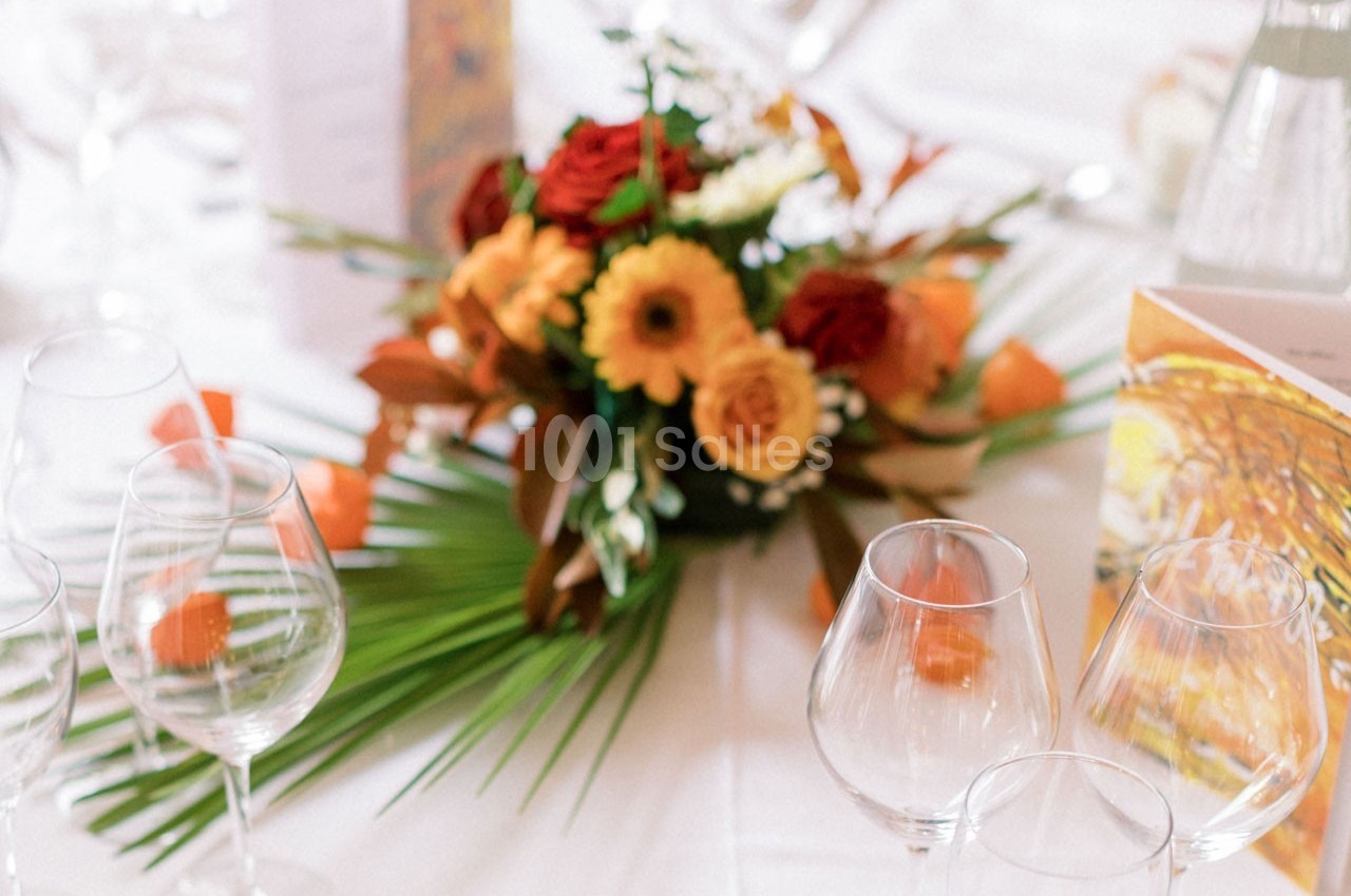 Centre de table floral avec des fleurs orange et rouges, entouré de verres à pied sur une nappe blanche.