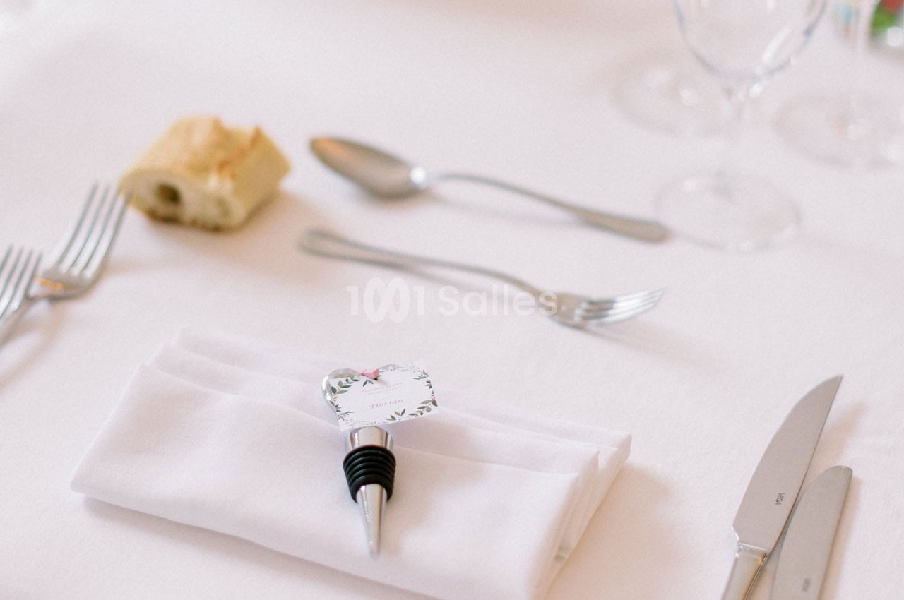Serviette pliée avec un bouchon décoratif, couverts disposés autour et morceau de pain sur une nappe blanche.