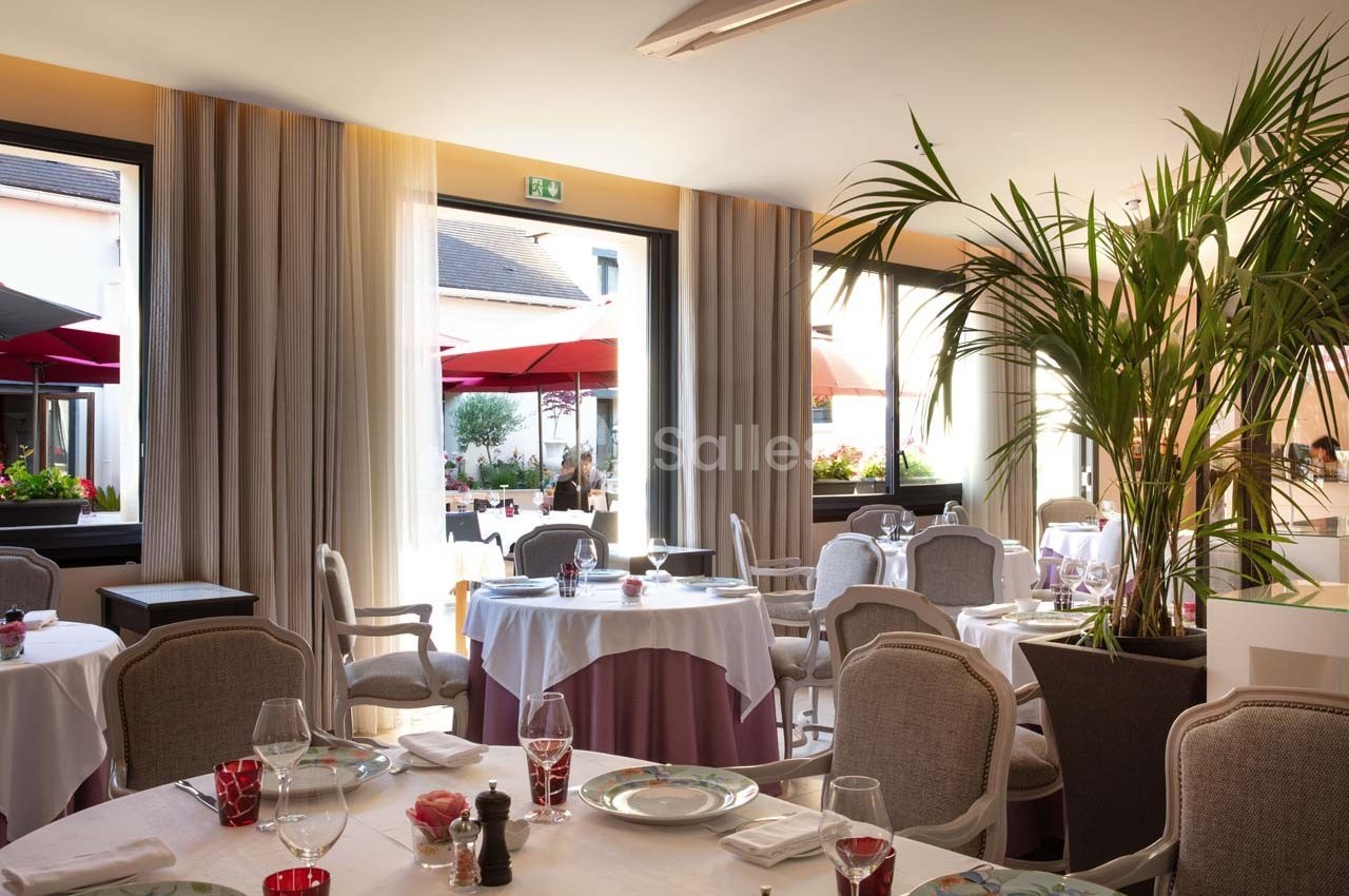 Salle de restaurant lumineuse avec tables dressées, grandes fenêtres ouvertes sur une terrasse avec parasols rouges.