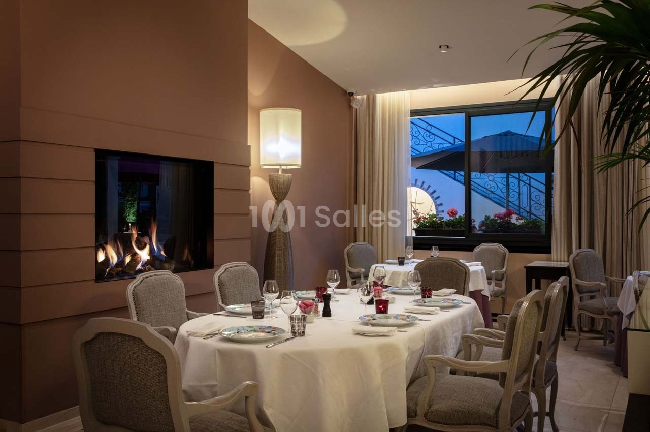 Salle de restaurant élégante avec tables dressées, cheminée allumée et vue sur une terrasse fleurie par une fenêtre.