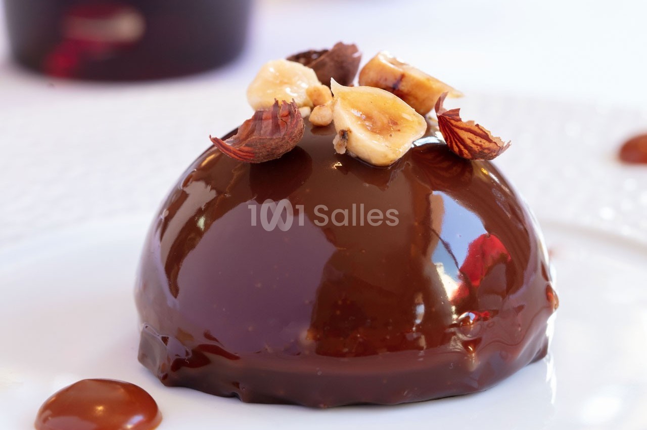 Dessert au glaçage chocolat brillant, garni de noisettes entières et concassées, présenté sur une assiette blanche.