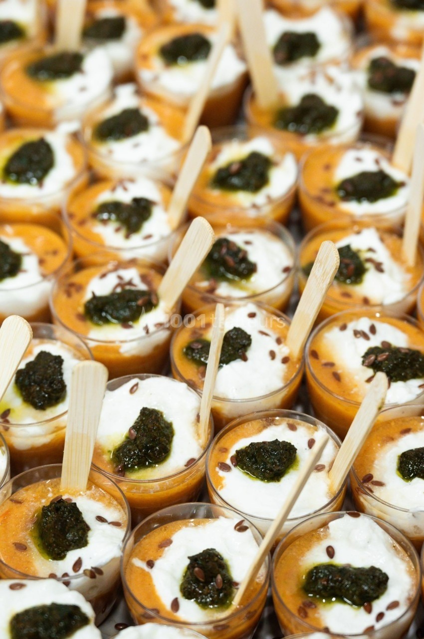 Verrines de soupe orange garnies de crème blanche, pesto vert et graines, avec des cuillères en bois.