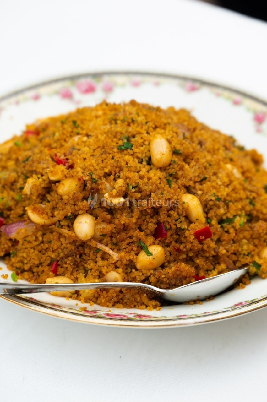 Assiette de couscous épicé garni de cacahuètes, herbes fraîches et morceaux de légumes, avec une cuillère en métal.