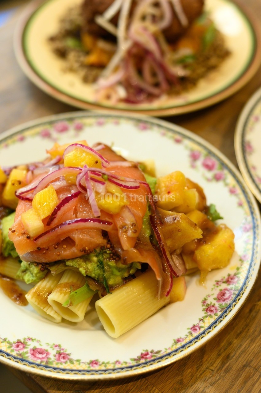Assiette de pâtes garnie de saumon fumé, mangue, avocat et oignons rouges, présentée sur une table en bois.
